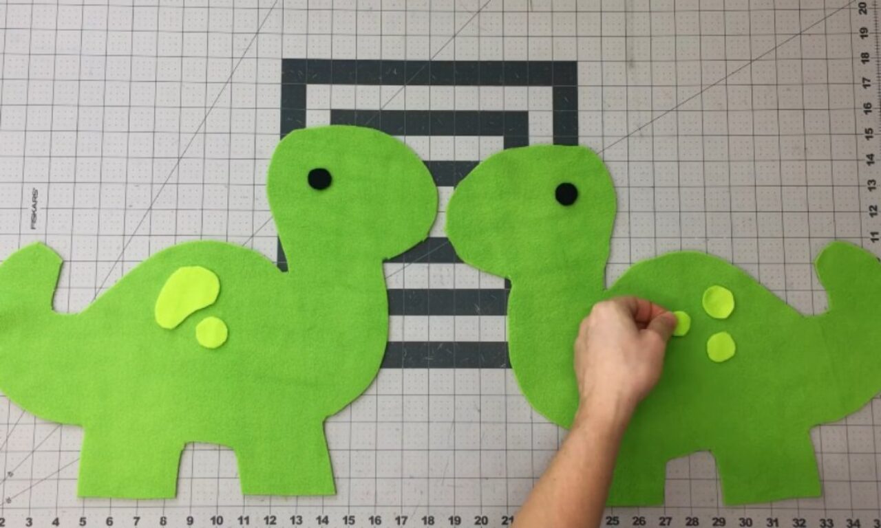 A person places light green felt spots onto the back of a bright green felt dinosaur cutout, lying next to another felt dinosaur on a gridded cutting mat.