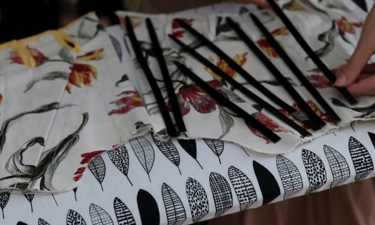 Several pieces of fabric with floral and leaf patterns lie on a table. Atop the floral fabric, which depicts stylized reddish-orange and grey-green tulips, are several thin black rods. Beneath the floral fabric is a piece of white fabric with a pattern of black and white stylized leaves. A person's hand is partially visible at the top right, holding one of the black rods