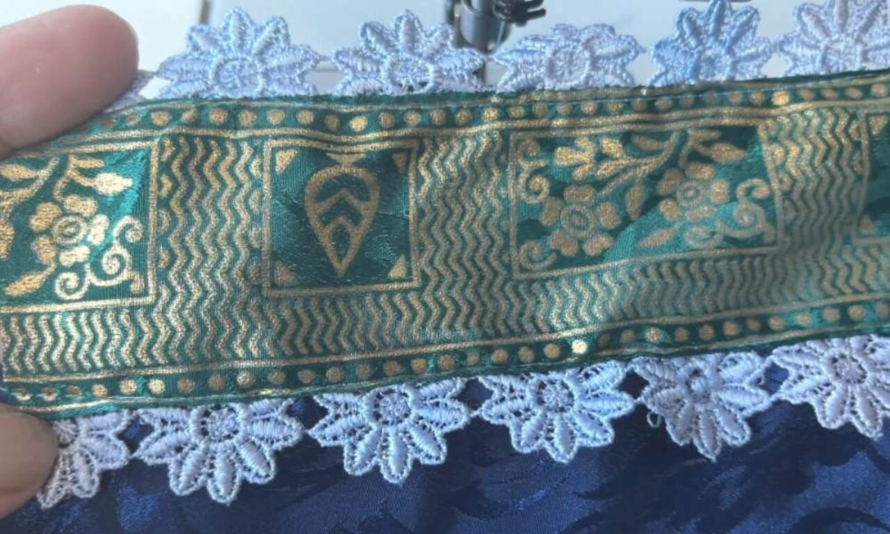 a dark blue fabric edge adorned with a white floral lace trim and an ornate green-and-gold ribbon featuring wavy patterns and stylized leaves and flowers.