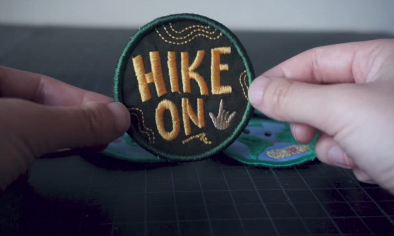 A circular embroidered patch with a dark background and green border being held between two hands. The patch features gold/yellow embroidered text reading "HIKE ON" and includes decorative elements like wavy lines and a small hand gesture. Another similar patch is partially visible beneath it.