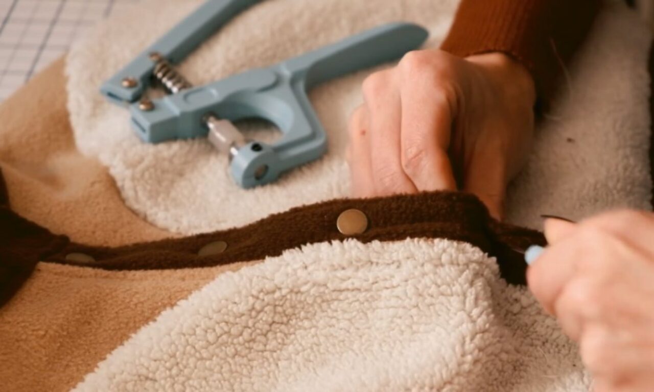 a hand using a light-blue snap tool to attach metal snap fasteners to a soft, two-toned fleece garment.