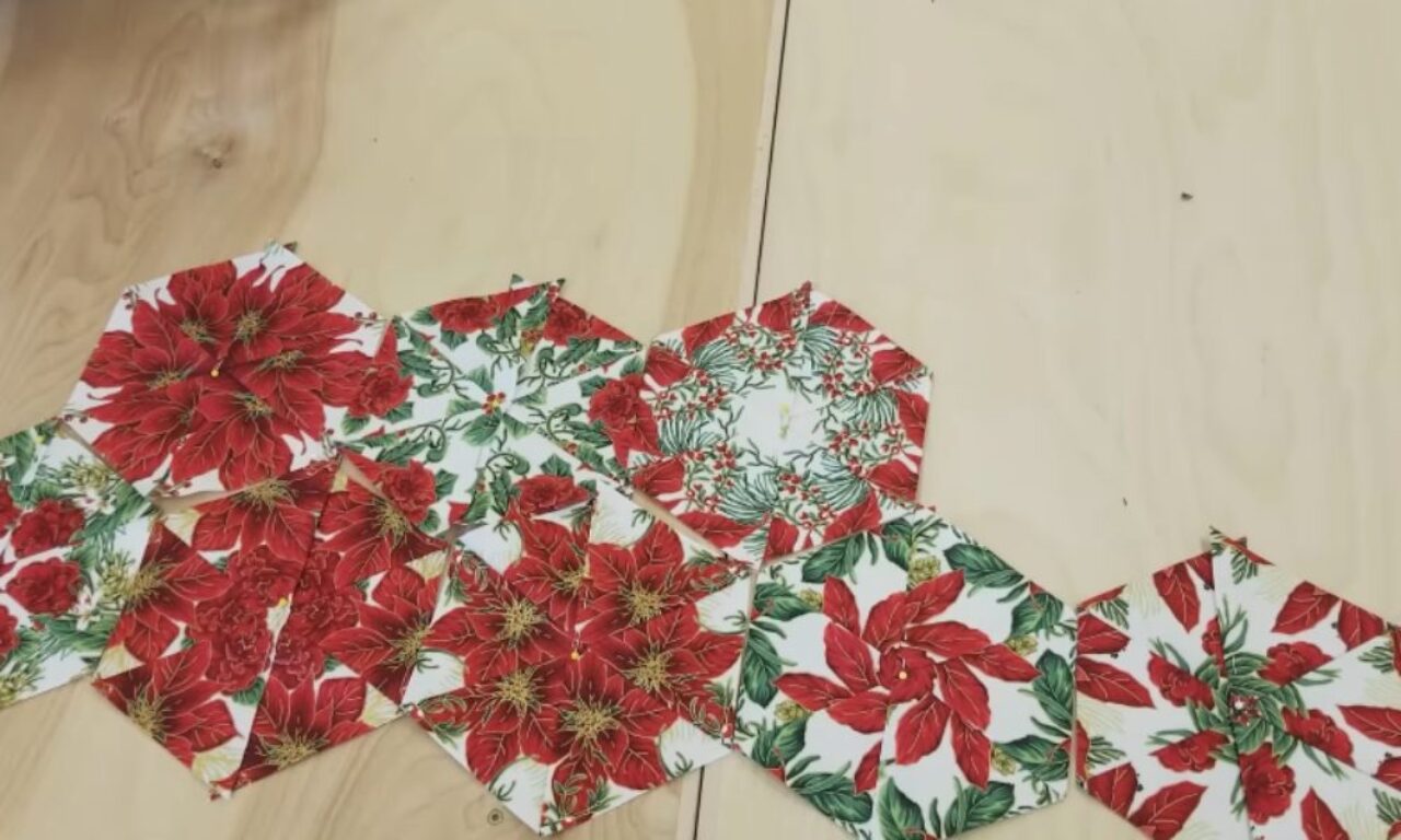 Hexagonal fabric pieces with Christmas patterns featuring poinsettias and holly laid out on a light-colored surface.