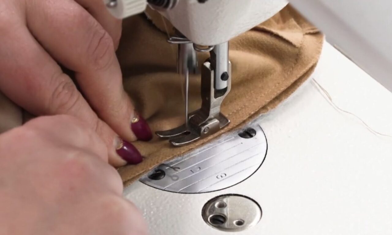 A close-up image of hands guiding fabric under the needle of a sewing machine. The machine's presser foot is positioned above the fabric, and the needle is lowered, ready to sew. The fabric being sewn is a tan color, and the sewing machine has a white body with a visible stitch plate.