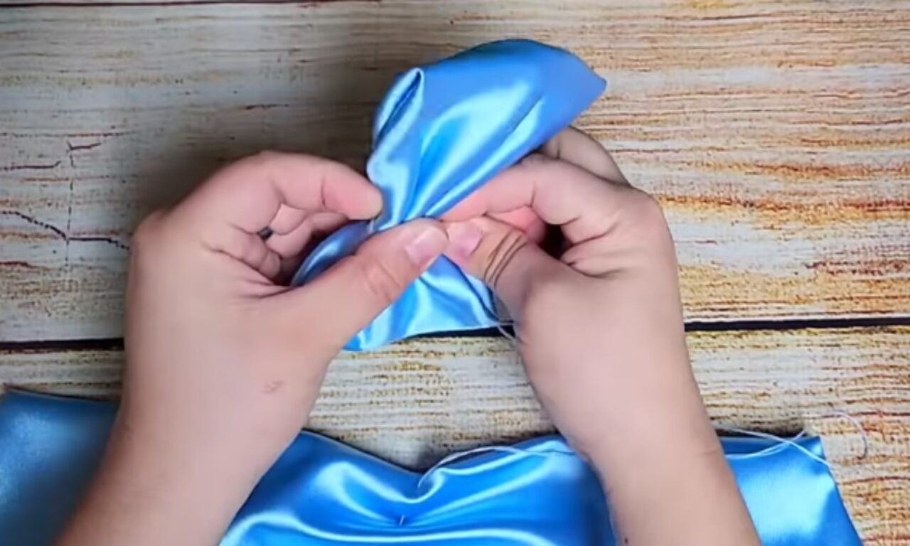 Hands holding and manipulating a piece of blue satin fabric, folding it into a bow shape on a wooden surface.
