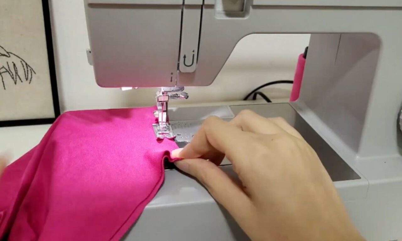 Close-up of a sewing machine stitching a piece of bright pink fabric. A hand guides the fabric under the needle.