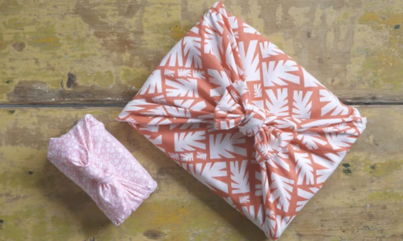 Two rectangular parcels wrapped in fabric using a knotted, furoshiki-style technique. One has a pink, subtle print, and the other features a white leaf pattern on a terracotta background. Both are resting on a worn wooden surface.