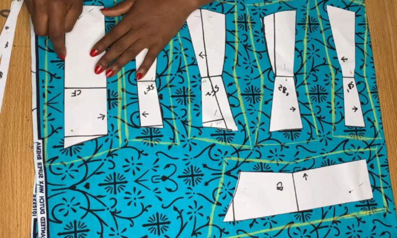 places paper sewing patterns onto a piece of bright blue fabric with a black floral design. The fabric is laid flat on a wooden surface. The paper patterns are labeled with letters and arrows, and yellow chalk outlines trace around them, indicating where the fabric will be cut. The scene suggests the beginning stages of garment construction.