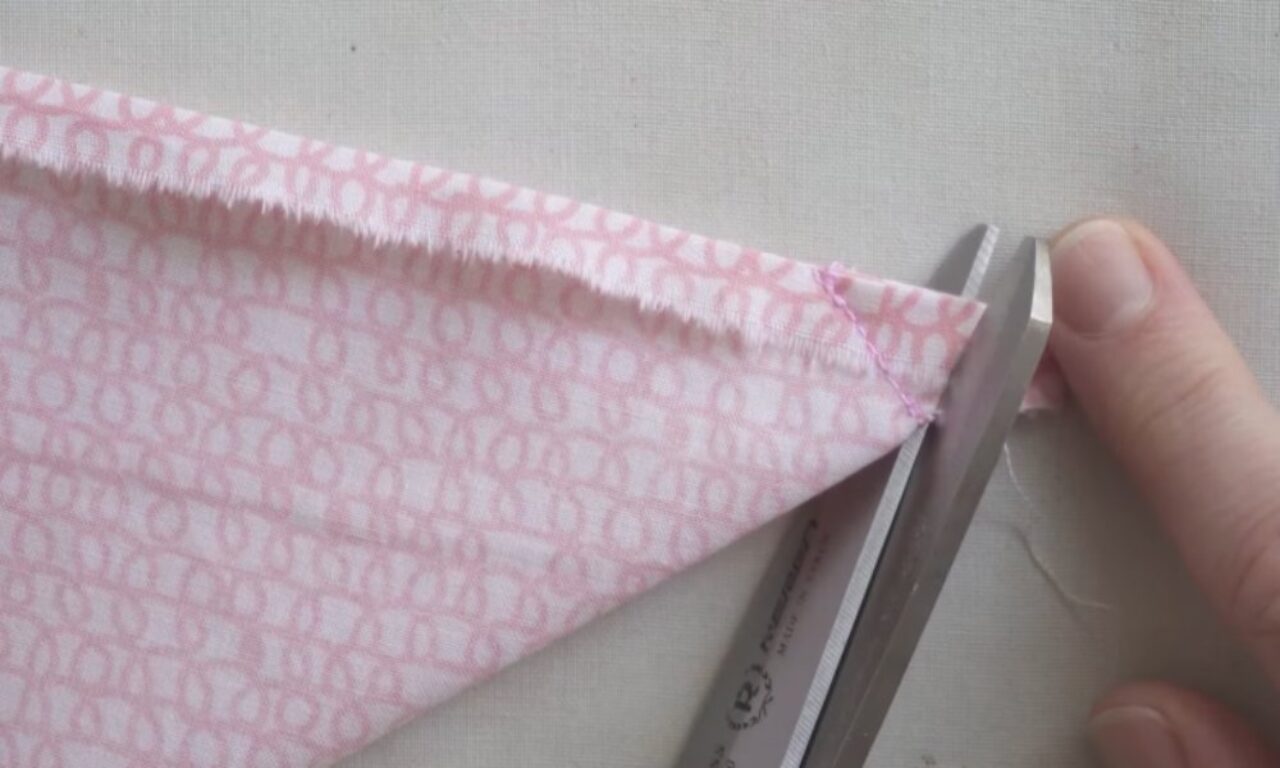 a pair of scissors cutting a pink fabric triangle with a patterned design. The fabric edges are frayed, and the cutting action is being performed with a hand holding the scissors near the fabric. The background is a light, neutral color.