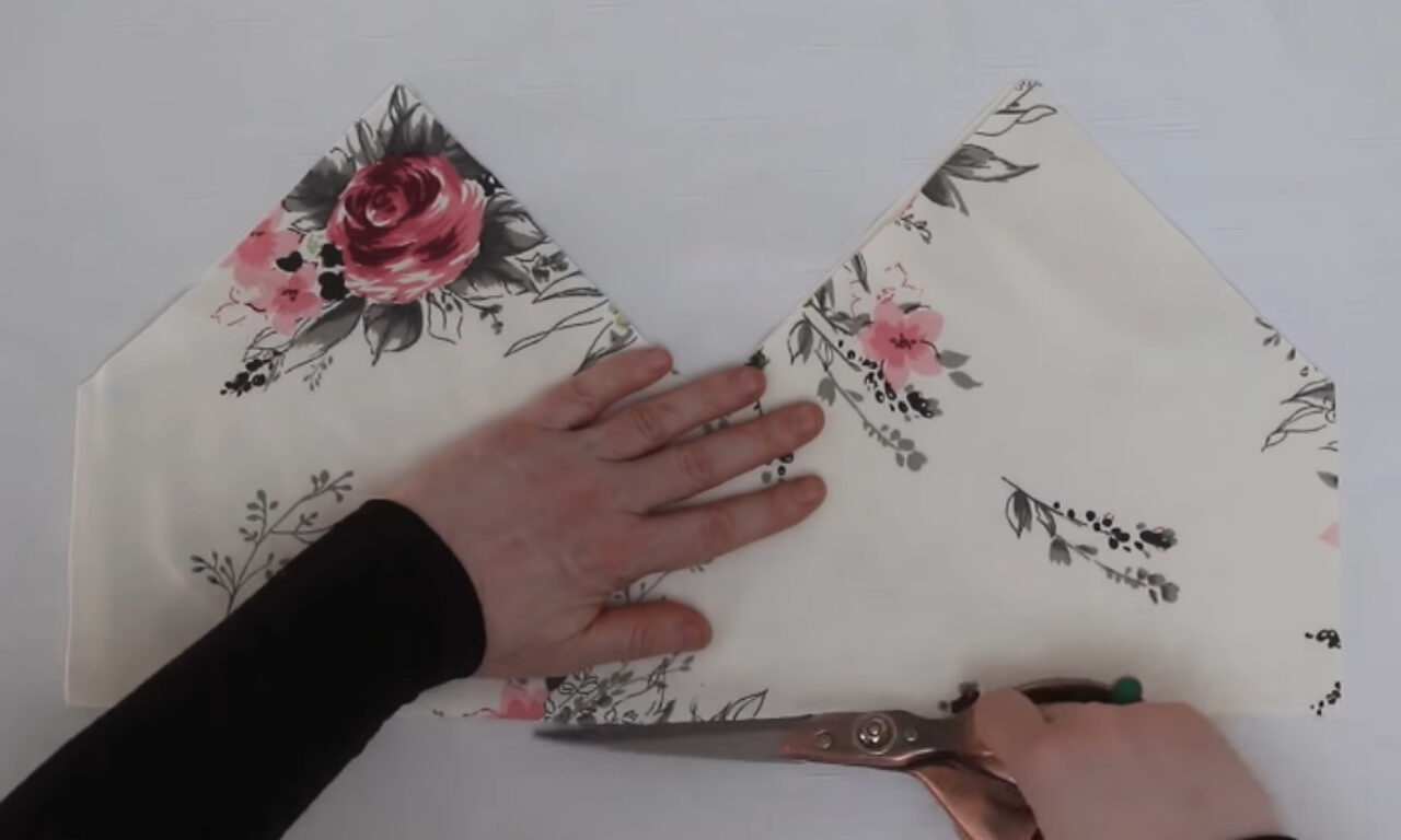 **A person's hands cut a piece of off-white floral fabric, patterned with pink and red roses and grey foliage, on a white surface. The fabric is folded to create two triangular points, and the person is using large, copper-handled scissors to cut along the bottom folded edge.**