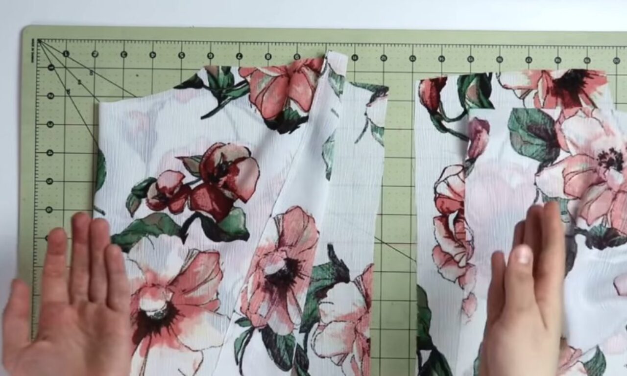 a white floral fabric with pink flowers and green leaves, laid flat on a green cutting mat. The fabric is divided in two down the middle, and two hands are placed on either side, smoothing out wrinkles.