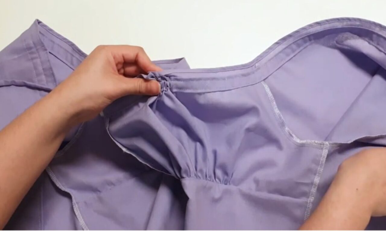 a person's hands adjusting the elasticized waistband of a lavender-colored garment, possibly pajama pants or shorts, against a plain white background.