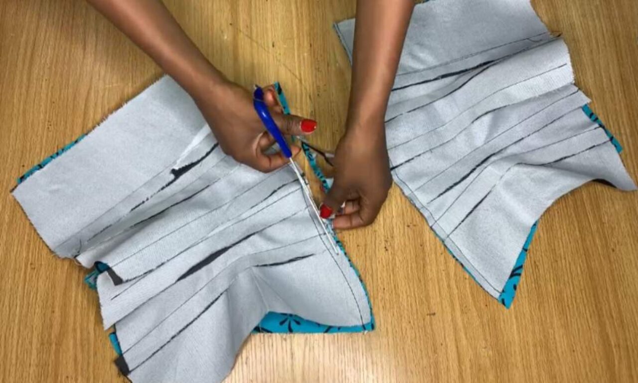 The person is holding the fabric down with their left hand and cutting it with their right hand, which is holding a pair of blue-handled scissors. The fabric is light gray with a blue patterned lining. It appears to be a skirt or dress pattern, as it has been cut into multiple pleats. The background is a light-colored wood surface.
