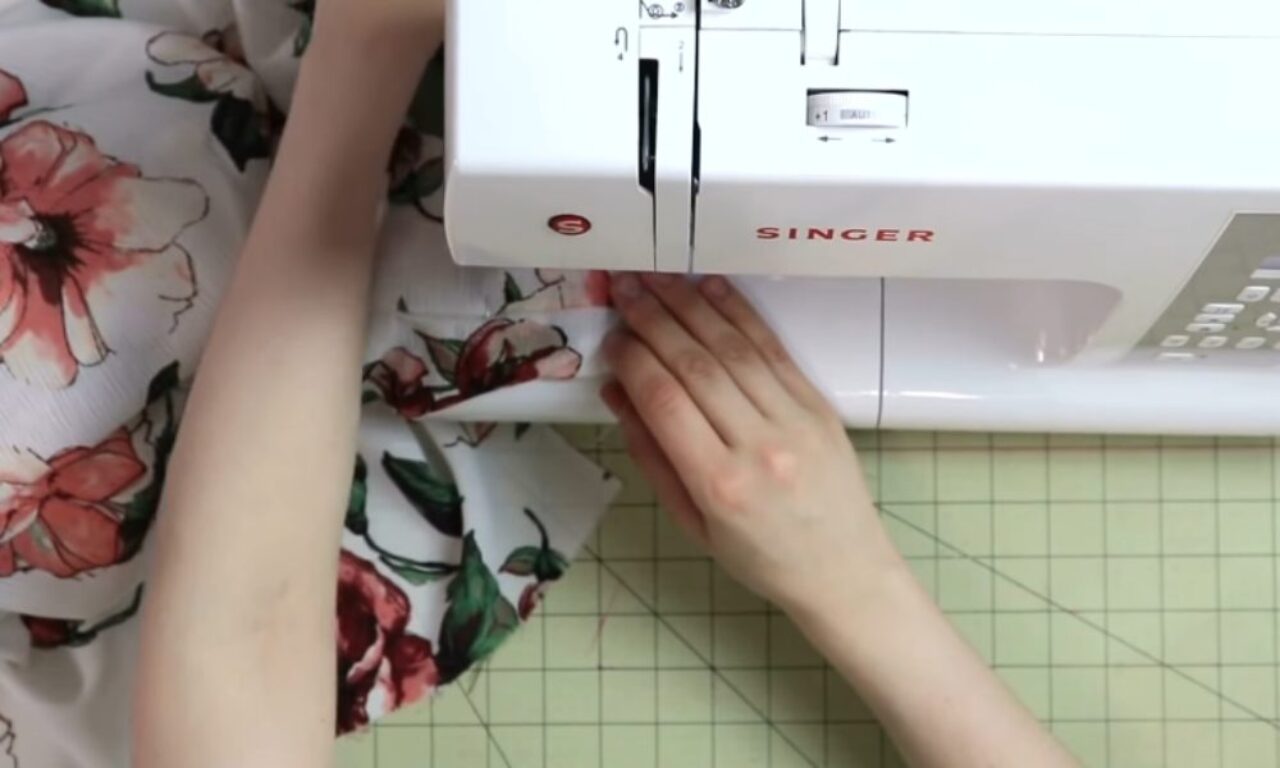 a person's hands guiding white fabric with a pink floral print through a white Singer sewing machine resting on a green gridded cutting mat.