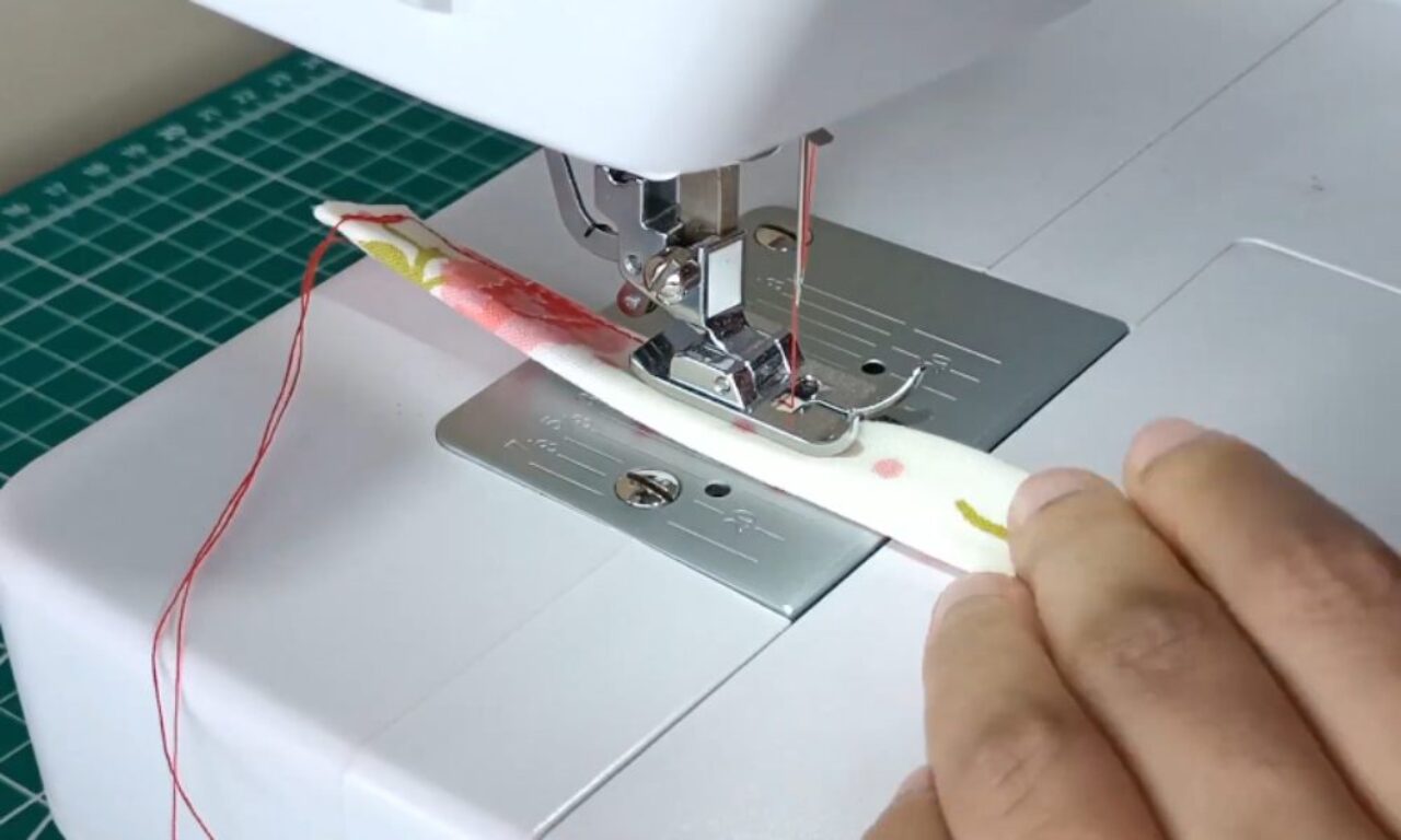 a sewing machine in action, showing the needle and presser foot area. A hand is guiding fabric with red and yellow markings through the machine. A green cutting mat is visible in the background.