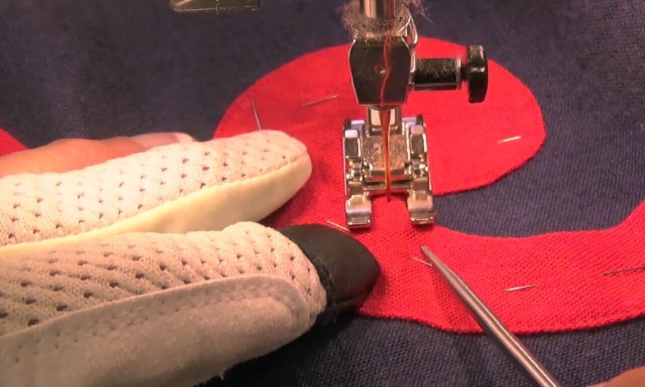 a gloved hand guiding a sewing machine needle as it stitches a red fabric shape pinned onto a dark blue background.