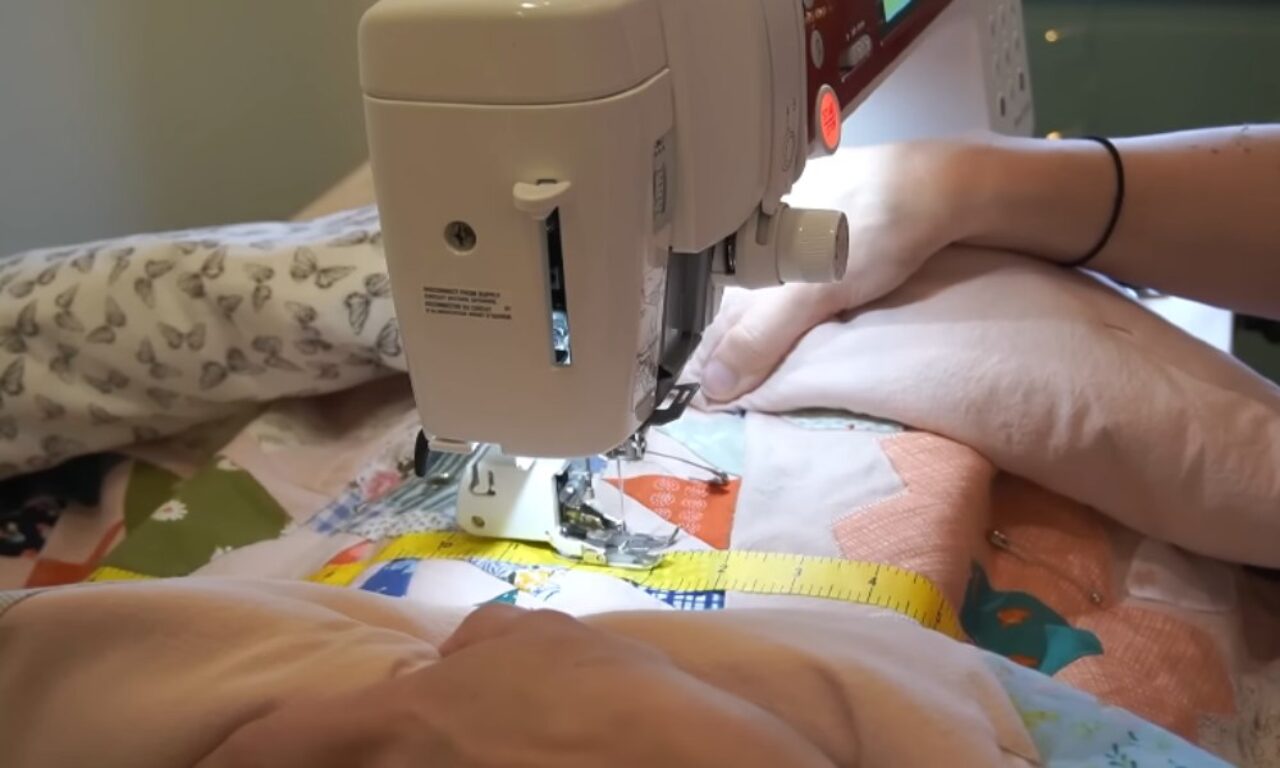 a sewing machine quilting a patchwork quilt. The machine is beige and the quilt is made of colorful squares, with a yellow measuring tape and safety pin visible on the fabric. The person's hands are visible, guiding the fabric under the needle.