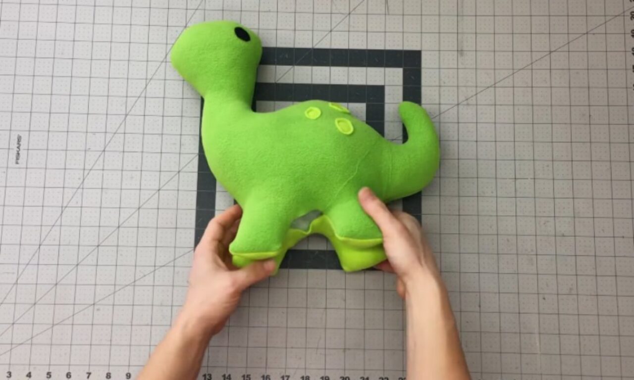 a bright green, plush dinosaur toy. The dinosaur has a simple design with small yellow buttons on its back and a black eye. It is placed on a gray cutting mat with grid lines for measurement. The background includes a white surface with faint grid markings.
