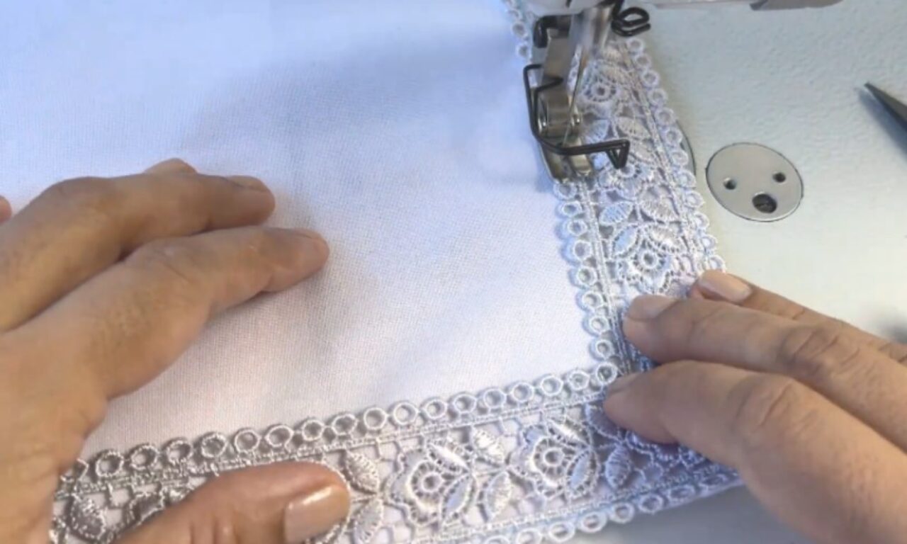 hands guiding white fabric and intricate white lace trim under the needle of a sewing machine.