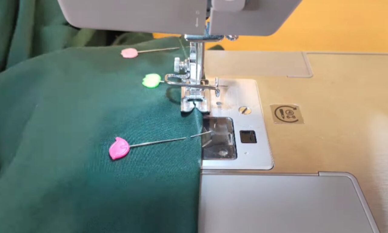 A sewing machine needle is stitching a seam on dark green fabric. Three sewing pins with colorful, flower-shaped heads are holding the fabric in place.