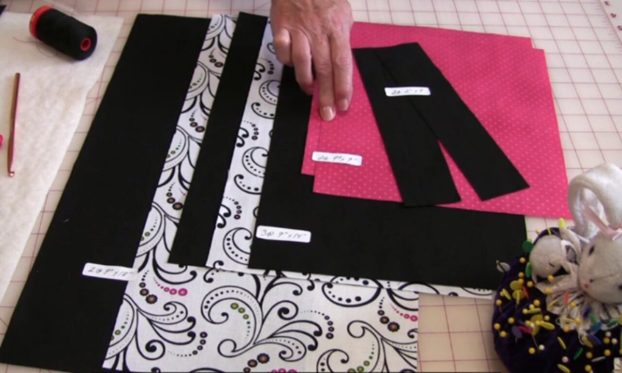 Assorted fabric pieces in black, white paisley, and pink with white dots are laid out on a cutting mat. Some pieces are labeled with dimensions. A hand is visible, along with a spool of black thread, a crochet hook, and a bunny-shaped pincushion.