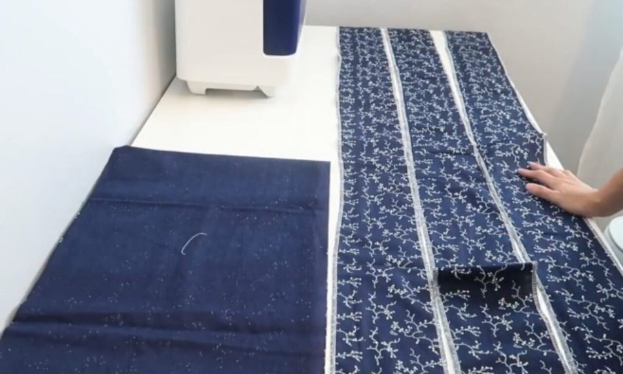 A sewing workspace with a white sewing machine in the background. On the table, there are two sets of dark blue fabric pieces: one solid and one with a white floral pattern. The patterned fabric is cut into several long strips, and a person’s hand is visible arranging the fabric on the right side of the table.