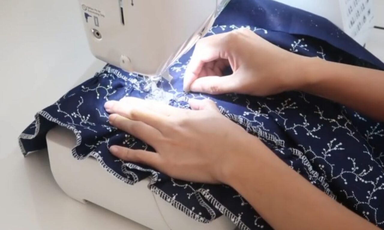 A person is using a sewing machine to stitch a piece of dark blue fabric with a white floral pattern. The hands are carefully guiding the fabric through the machine, ensuring precise stitching. The sewing machine is white and appears to be in good condition, placed on a light-colored surface.