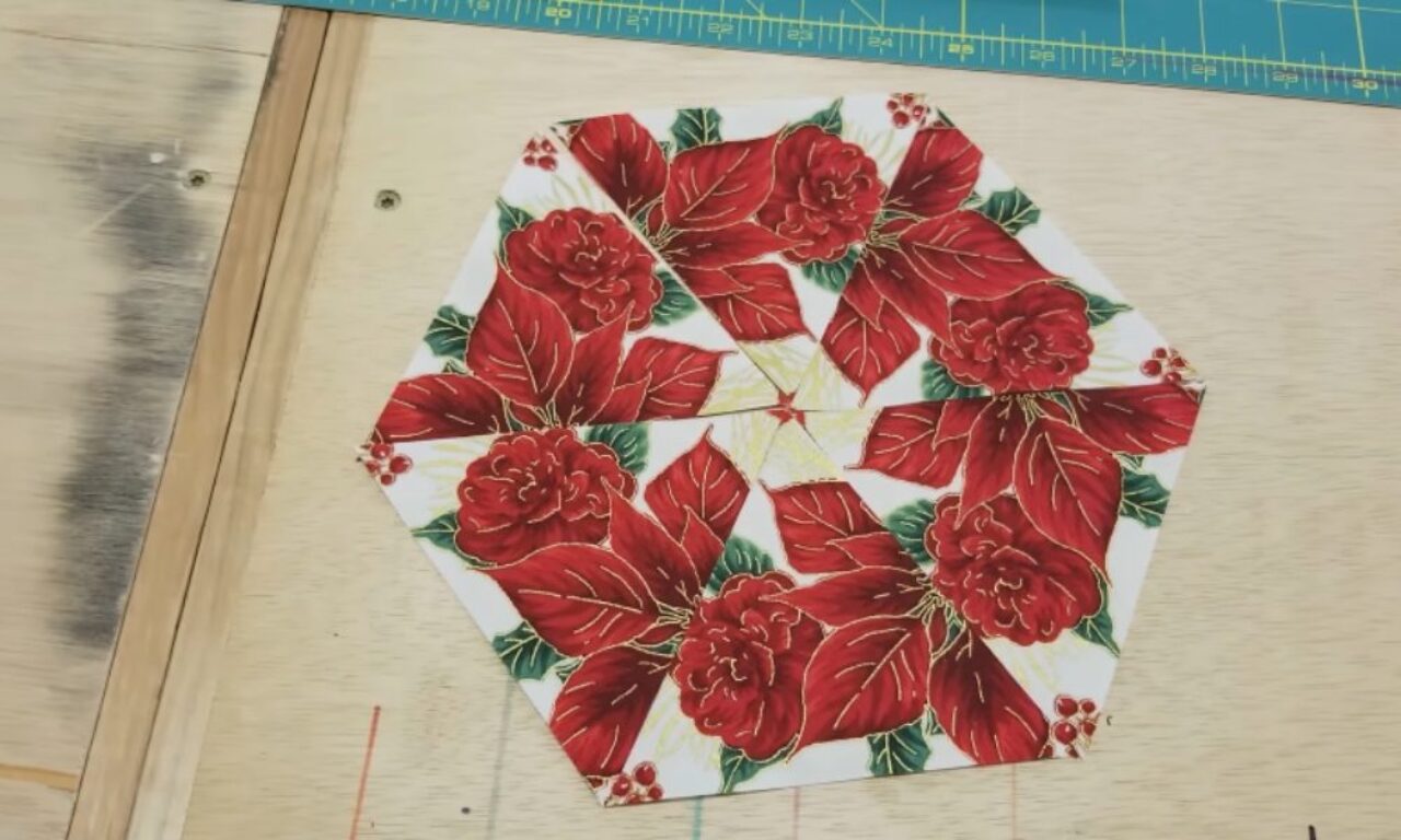 Hexagonal quilt block featuring a pattern of red poinsettia flowers with green leaves and small red berries on a white background, placed on a wooden surface with a blue measuring ruler at the top.