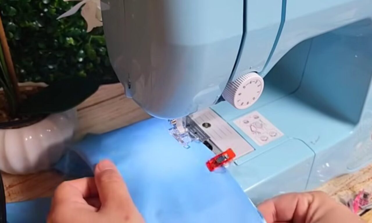 a light blue sewing machine with a person's hands guiding light blue fabric underneath the needle. The machine sits on a wooden table, with a potted plant and other items visible in the background.