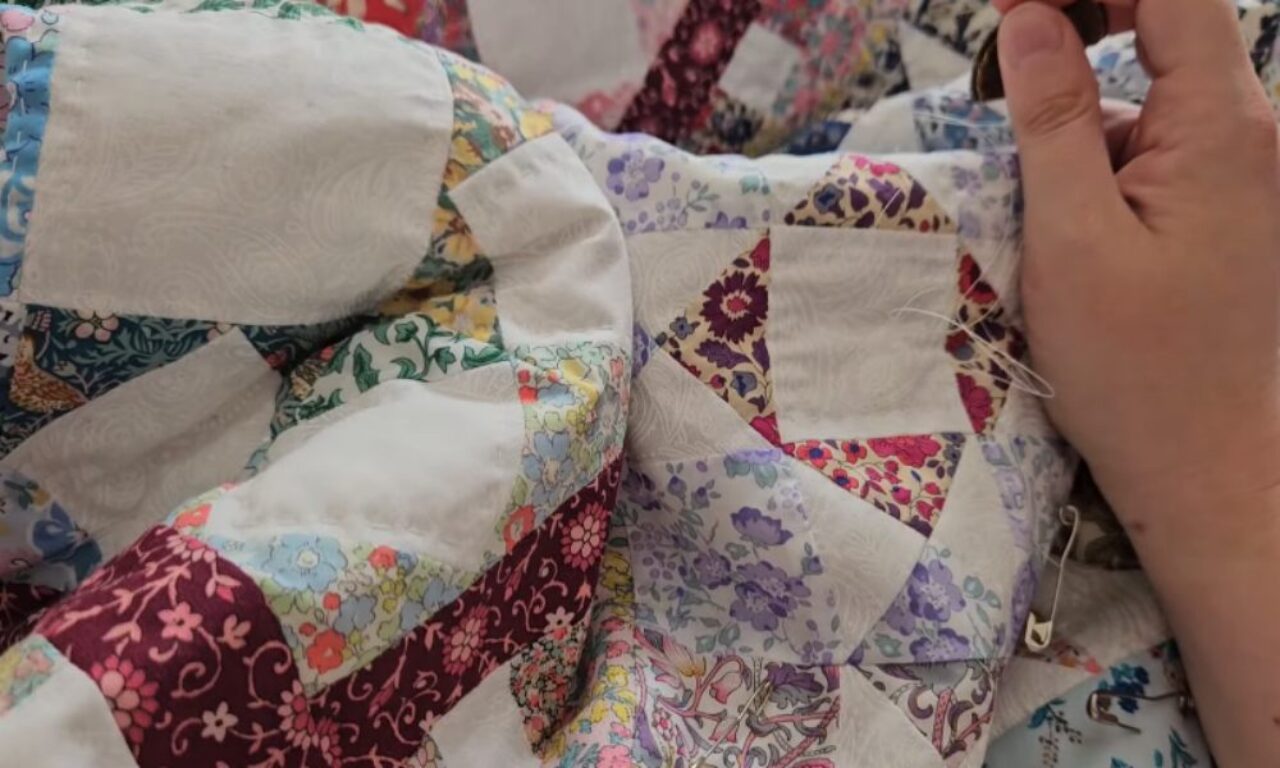 hand-stitching a colorful patchwork quilt made of various patterned fabrics in shades of red, purple, blue, green, and white. The quilt features geometric shapes and floral designs, and a safety pin is visible securing part of the fabric.