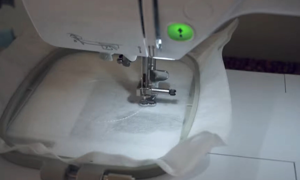 Close-up of an embroidery machine needle and presser foot stitching white fabric held in a hoop.