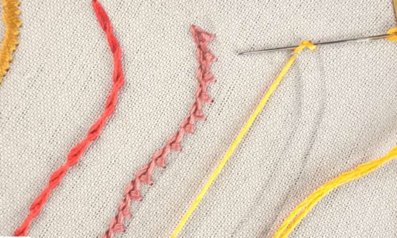 embroidery on beige fabric showing three different stitches. On the left is a red twisted chain stitch, in the center is a pink crested chain stitch, and on the right, a yellow thread is being used with a needle to create another stitch. The needle is partially inserted into the fabric.