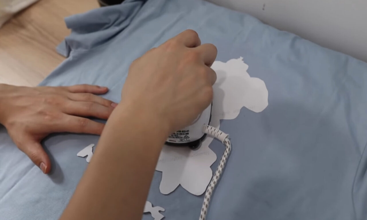 A person is using a white iron to press a white cut-out design onto a light blue T-shirt. One hand holds the fabric steady while the other applies pressure with the iron. The T-shirt is laid flat on a wooden surface, and the background features a white wall.