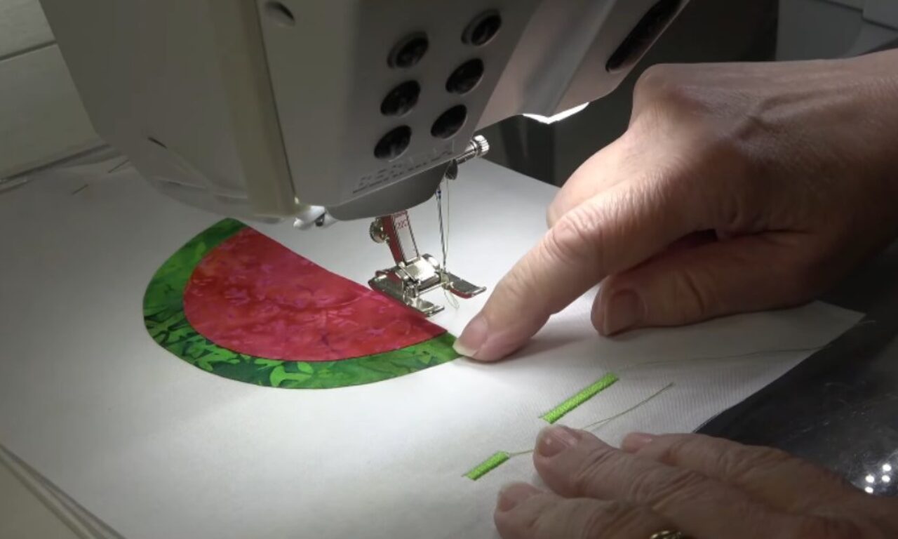hands guiding fabric under a sewing machine needle, stitching the green rind of a watermelon slice appliqué made from red and green patterned fabric onto a white base material.