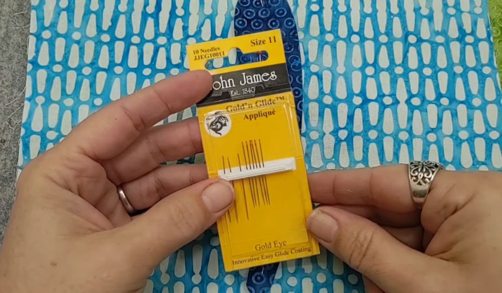 A pair of hands holding a yellow package of John James Gold'n Glide Applique needles, size 11, with a blue and white patterned fabric in the background.