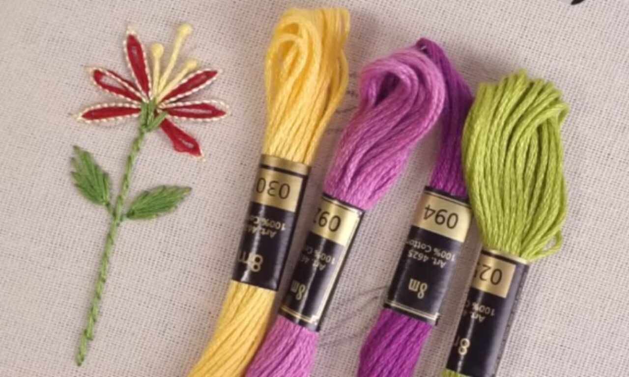 An embroidered red, yellow, and green flower on beige fabric next to three skeins of embroidery floss in yellow (0630), purple (0660), and light green (0620).