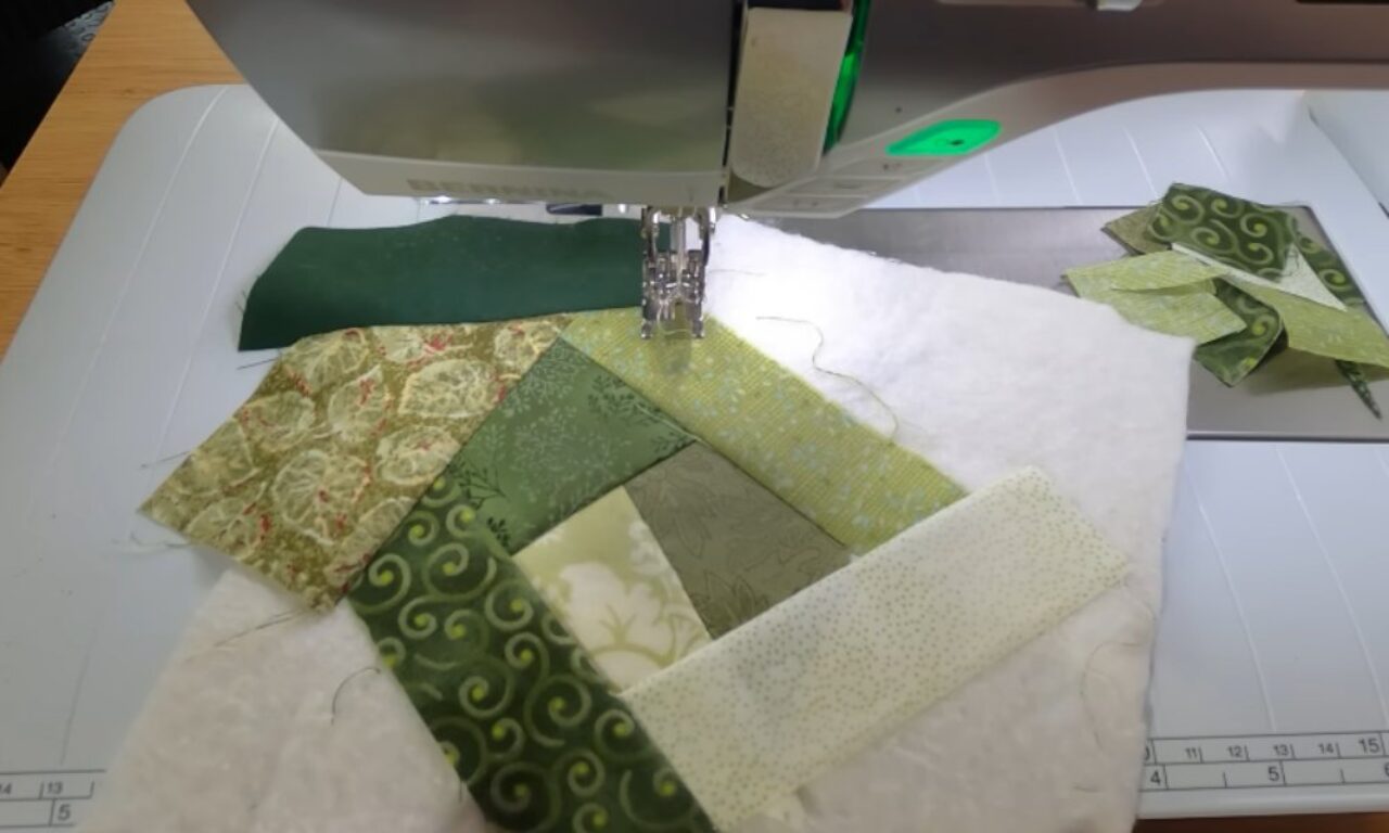 a sewing machine stitching together various shades of green fabric patches arranged in a square pattern on top of white batting, with additional fabric pieces and a cutting mat visible in the background.