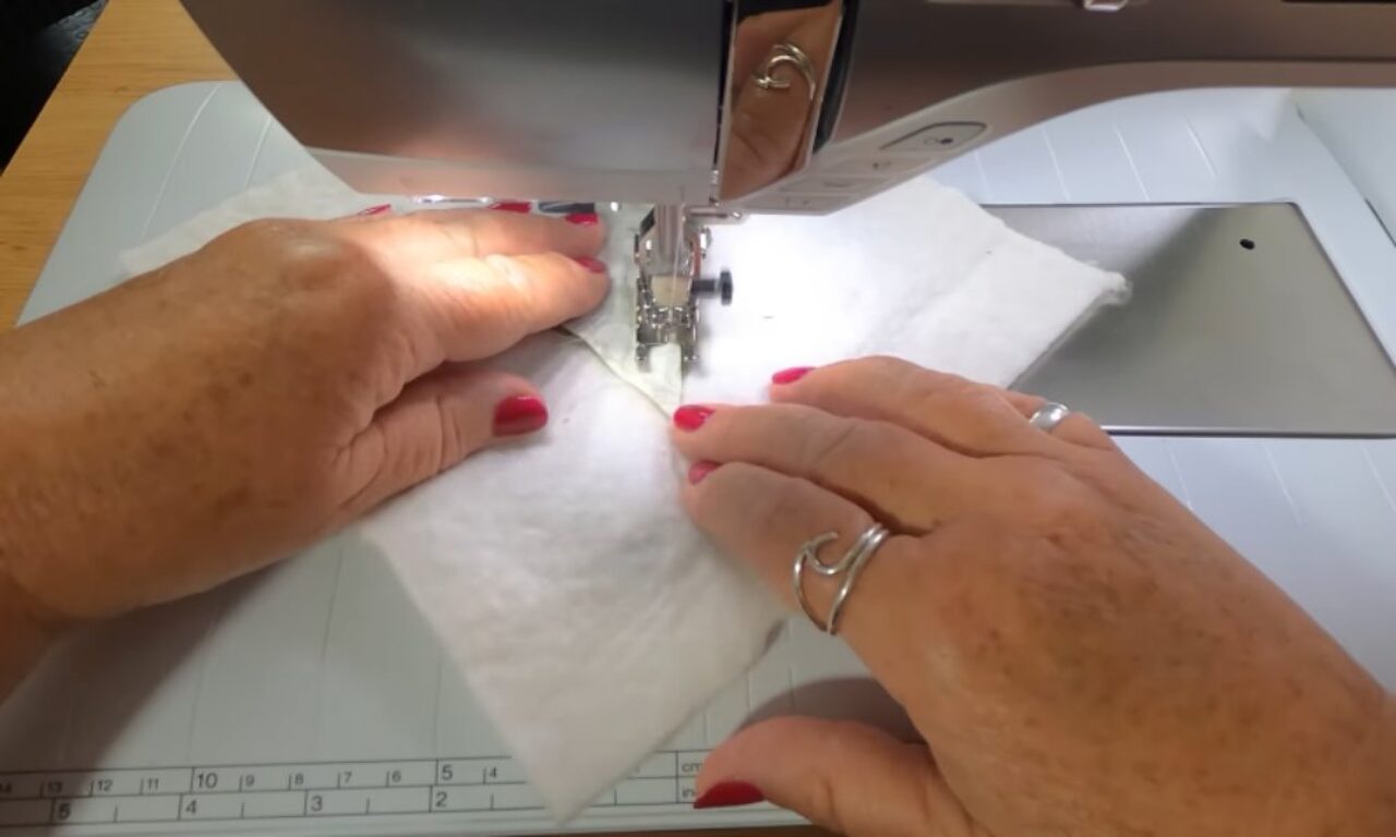 guiding white fabric through a sewing machine, stitching along a seam. The hands are positioned carefully, showing precise control during the sewing process. The machine is set on a white surface with measurement markings.