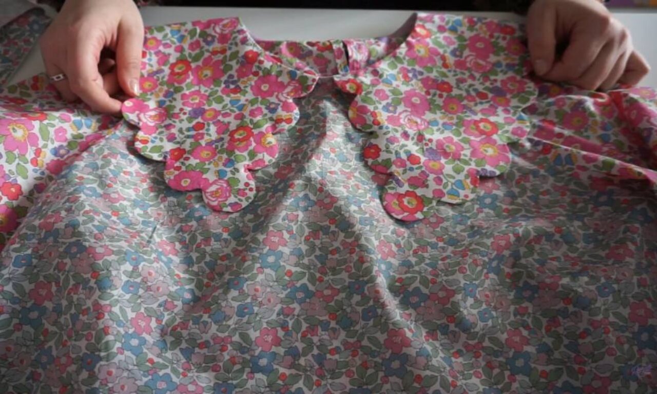 hands holds a child's dress sewn from two coordinating liberty-style floral prints, with a scalloped collar in bolder pink flowers over a softer pastel flower pattern on the body.