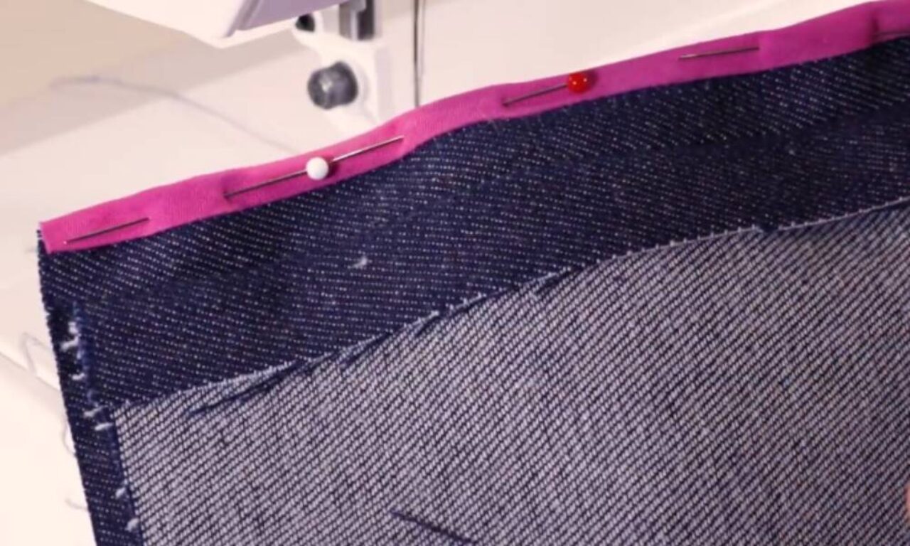 Sewing project in progress: denim fabric with a pink trim edge being pinned before sewing on a sewing machine.