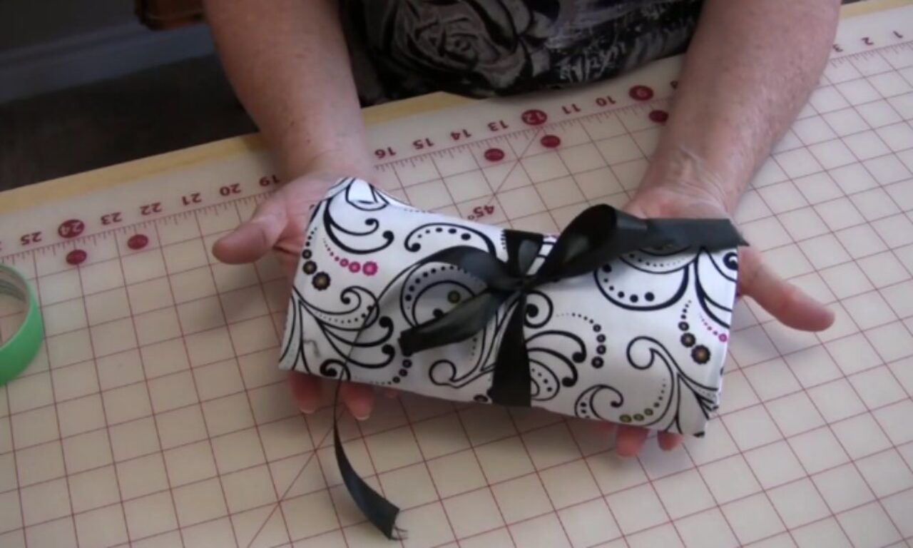 Hands hold a small item wrapped in white fabric with black swirls and pink dots, tied with a black ribbon, on a pink crafting mat.