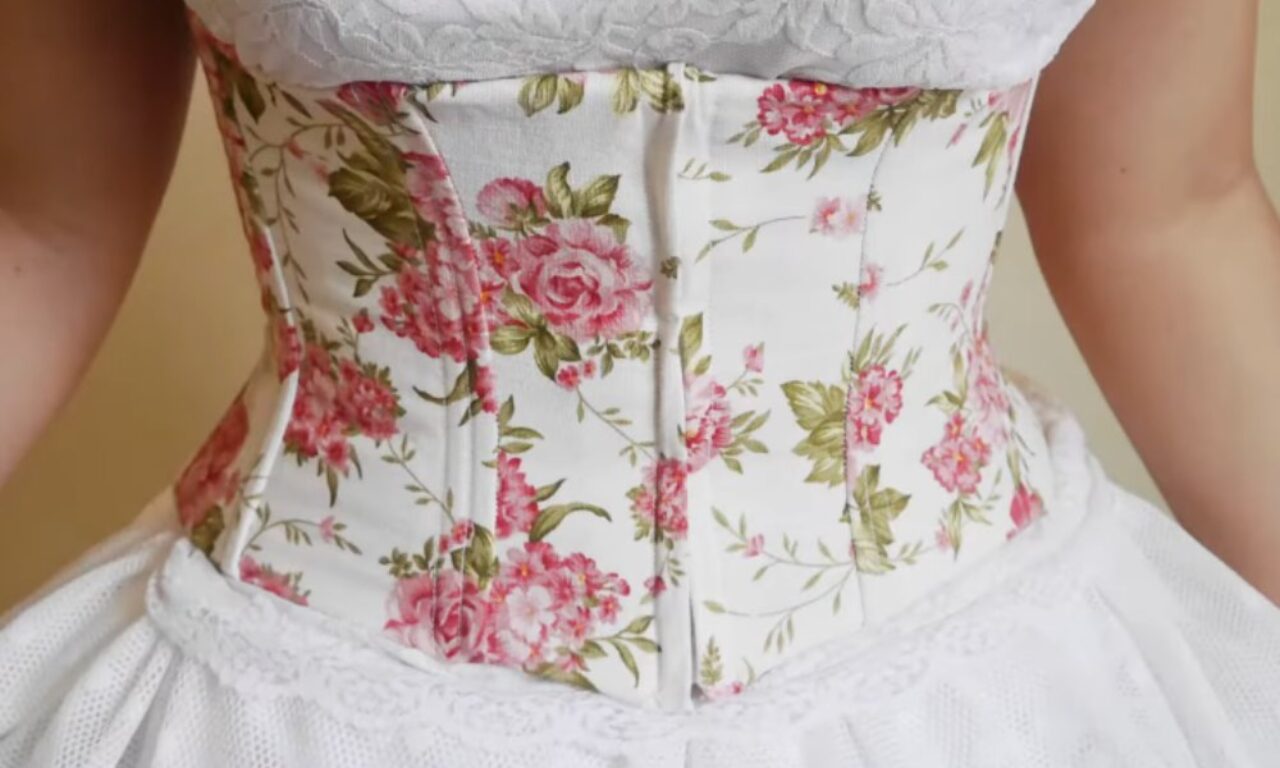 a corset or bodice with a white background and pink floral print pattern featuring roses and green leaves. The garment appears to be part of a dress or wedding gown, with white lace or fabric visible at the top and bottom edges. The corset shows structured boning and a fitted waistline.