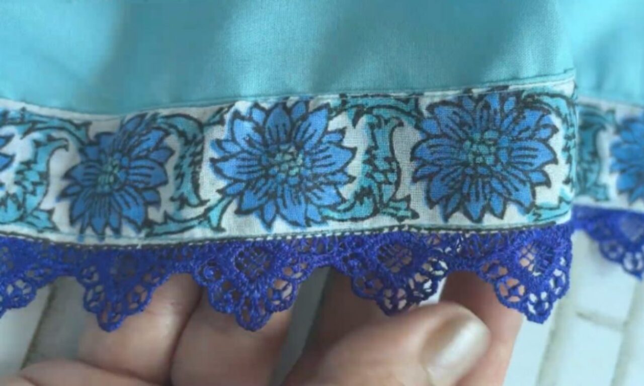 showing the hem of a light blue garment, detailed with a white band featuring a blue floral pattern and finished with a dark blue lace edge. Fingers hold the hem from below.