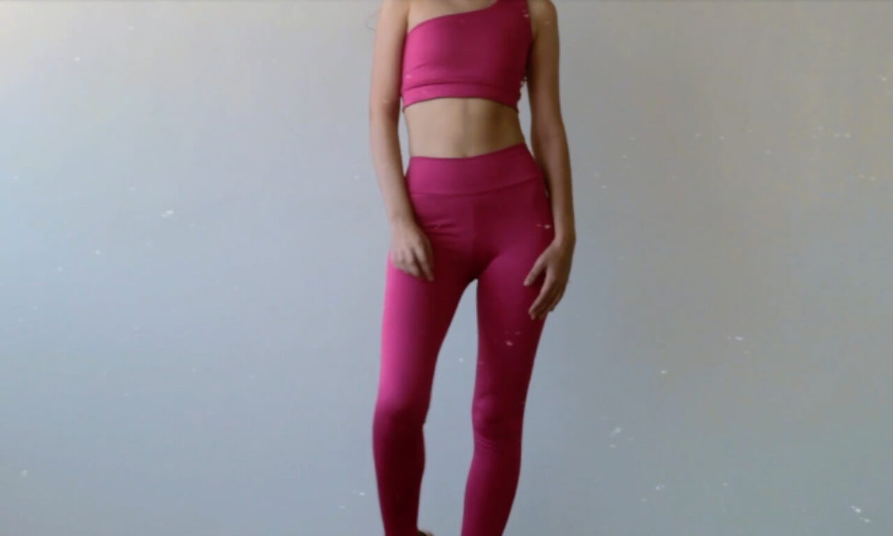 A person standing against a plain light background, wearing a one-shoulder pink sports bra and matching pink leggings. Their face is out of frame, and they are posed with one hand resting by their side.