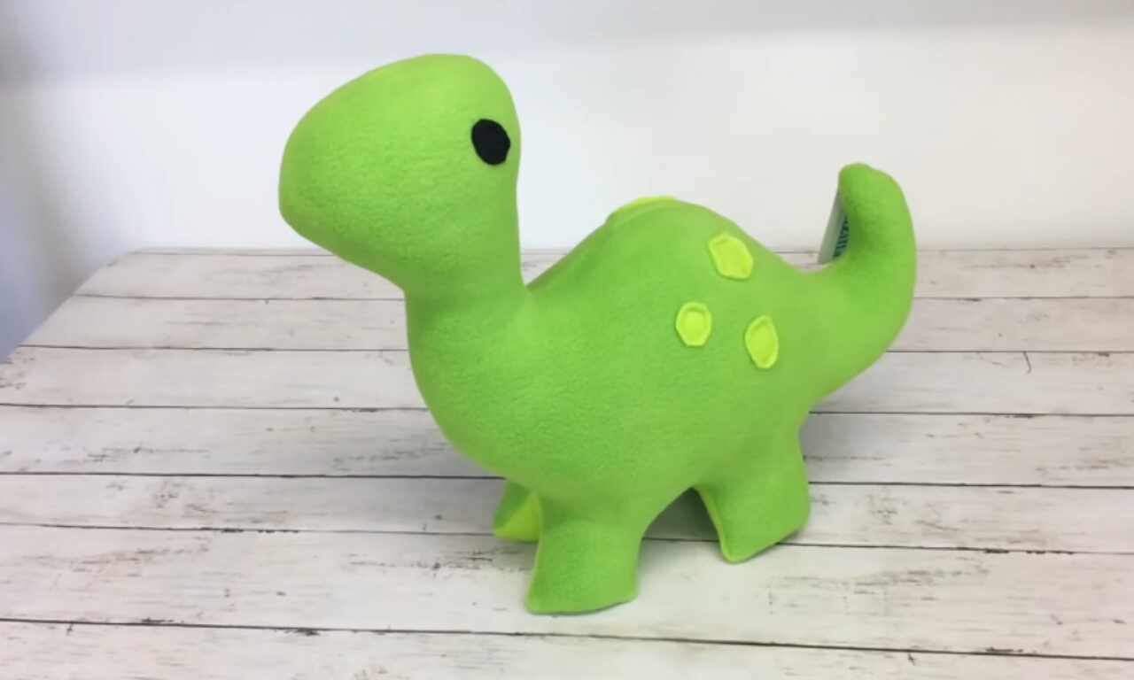 A bright green, soft, plush dinosaur toy with simple black eyes and lighter green spots on its back, standing on a surface that looks like light-colored wood planks.