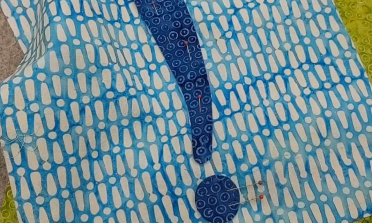 Fabric pieces pinned together to form an exclamation point shape. The layered fabric is dark blue with a circular pattern, and the background fabric is blue and white with an abstract pattern.
