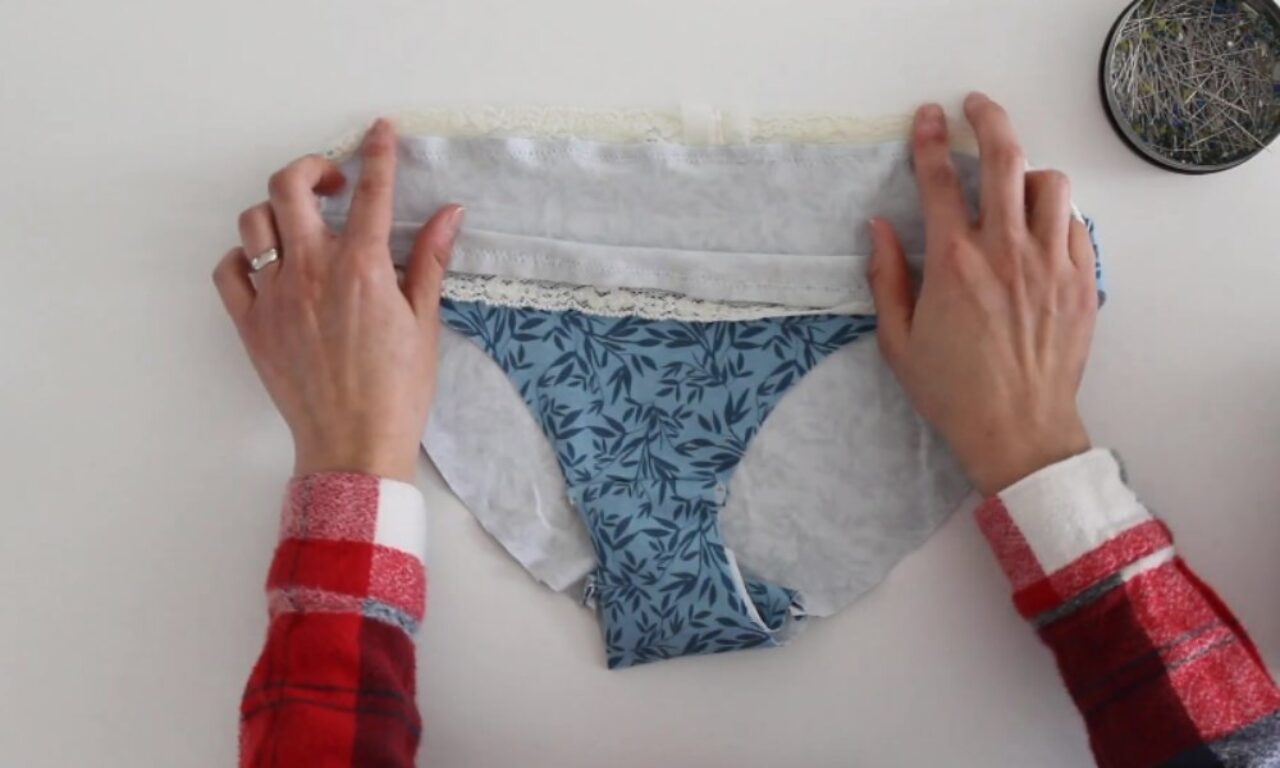 Hands hold open the waistband of blue underwear with a leafy pattern, revealing a grey lining and white lace trim. A pin cushion is in the top right corner.