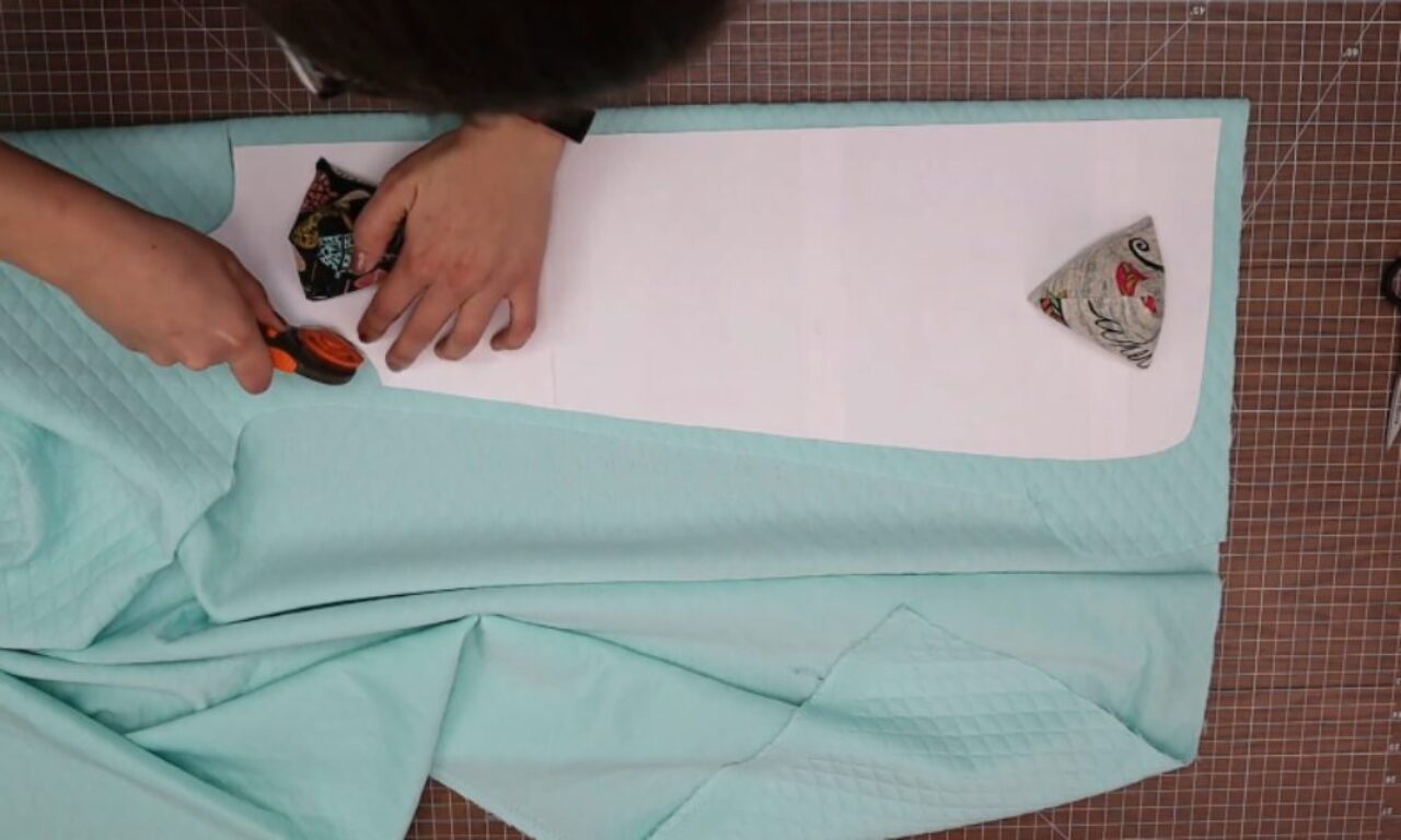 A person is cutting a curved shape out of a piece of light blue fabric using scissors, with a white paper template underneath. Two small fabric triangles, one black and one gray with a floral pattern, are placed on the template.