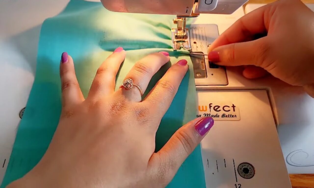 A person sewing a piece of turquoise fabric on a sewing machine, with their left hand holding the fabric steady and wearing a ring with a floral design, while their right hand guides the fabric through the machine. The sewing machine has the brand name "Effect" visible on it.