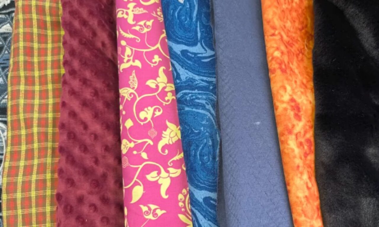 An image of folded fabrics with various patterns and colors, including red and yellow plaid, maroon with raised dots, pink with yellow floral designs, blue and white swirls, solid blue, orange and yellow swirls, and solid black.