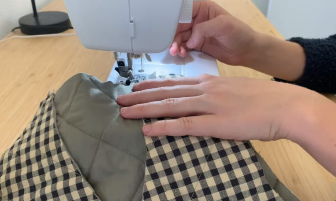 Hands operating a sewing machine, working with a black and white gingham fabric paired with a solid gray material. The machine sits on a wooden table surface, and the hands are guiding the fabric through the needle area.